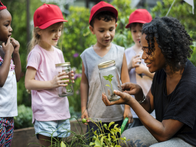 Afro American primary school teacher supporting experiential learning  through outdoor education holding a jar with a plant and showing it to students 