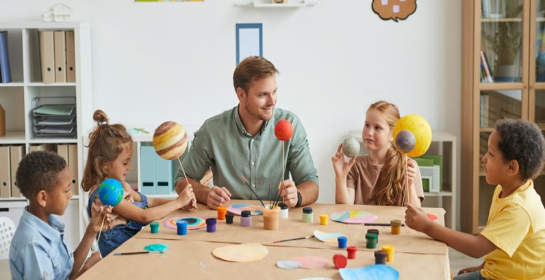 male caucasian teacher in the classroom with group of children at the table offering experiential learning activities exploring planets, arts & crafts 